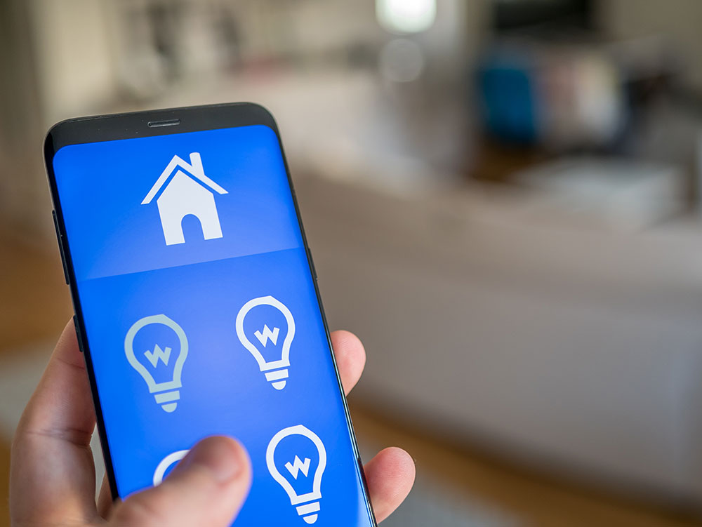 Nærbildet av en telefon som viser et stort blått bildet med symbol av et hus og fire lyspærer, og dette er appen Dali lysstyring fra Micro Matic er kompatibelt med KNX og gjør dine nåværende lys- og bryterløsninger heldigitale