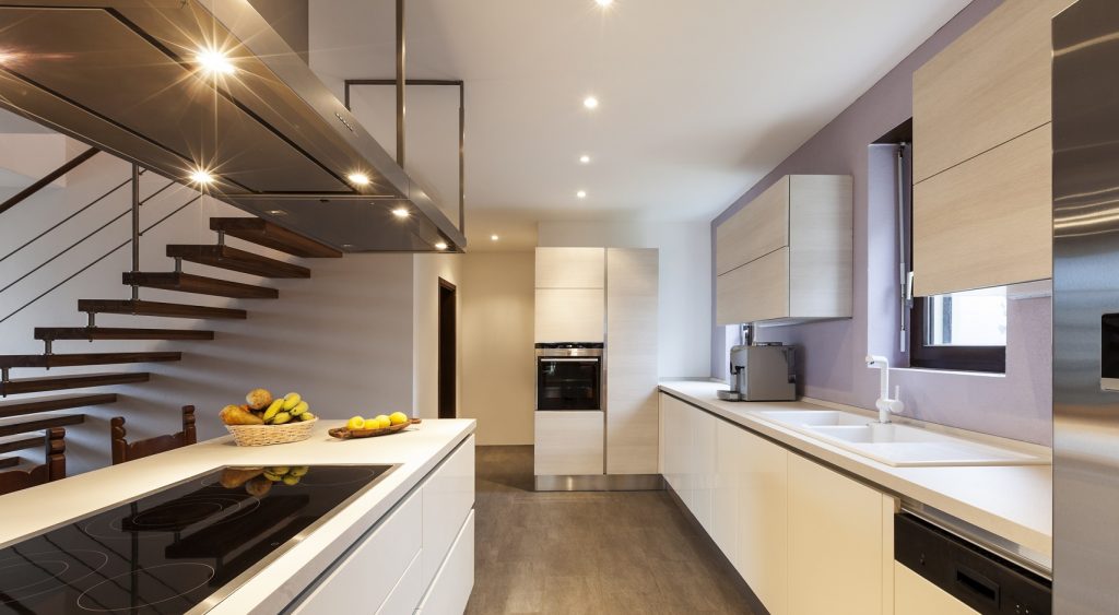 Et hvitt kjøkken med skap langs den ene veggen og en kjøkkenøy med koketopp og fin spottbelysning i den store viften over. Spotter lyser opp gjennom kjøkkenet.